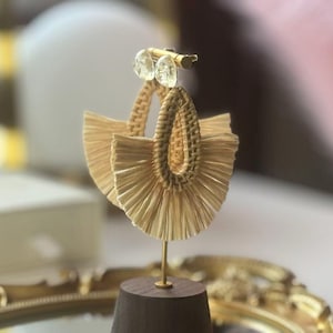 Peut inclure: Une paire de boucles d'oreilles en raphia beige tressé avec un motif en forme d'éventail. Les boucles d'oreilles sont présentées sur un support doré avec une base en bois. Des accents ronds et transparents sont au sommet.