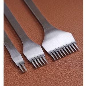 Pode incluir: Três ferramentas de trabalho em couro em aço inoxidável com pontas dentadas, dispostas do menor para o maior. As ferramentas estão sobre uma superfície de couro castanho, mostrando seu design afiado e preciso.
