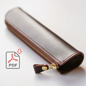 Könnte beinhalten: Eine braune Lederstiftetui mit einem goldenen Reißverschluss. Der Reißverschluss hat einen Lederzug aus braunem Leder.