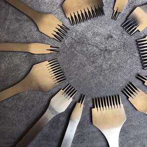 Può includere: Un set di dieci strumenti per l'artigianato del cuoio color oro con punzoni per cuciture neri. Gli strumenti sono disposti in cerchio su una superficie grigia testurizzata.