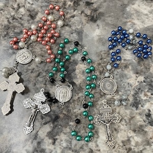 May include: Three rosaries with pearl beads in pink, green, and blue. Each rosary features a crucifix and a medallion with religious imagery. The pink rosary has white and pink beads, the green has black and green beads, and the blue has white and blue beads.