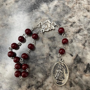 May include: A rosary with deep red beads and a silver-toned clasp. The rosary features a decorative clasp and a silver oval pendant with the words "INFANT OF PRAGUE".