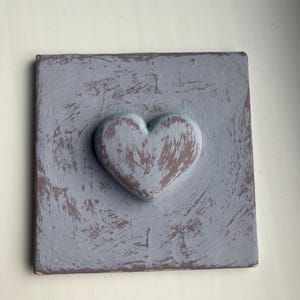 Könnte beinhalten: Ein quadratisches, dekoratives Holzstück mit einem erhabenen Herzdesign. Das Herz ist in einem gedämpften, staubigen Rosaton bemalt, und die quadratische Basis ist ein verwittertes Grau-Blau. Das Stück hat eine rustikale, handgefertigte Ästhetik.