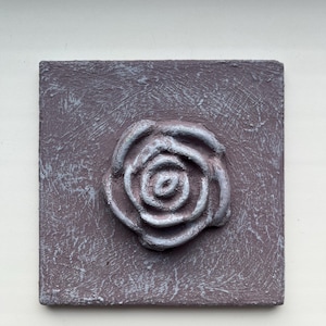 Puede incluir: Una pieza de arte mural cuadrada y texturizada, de color malva, con una rosa plateada en relieve en el centro. La rosa tiene pétalos detallados y es el punto focal de la obra de arte. El fondo tiene una apariencia desgastada y envejecida.