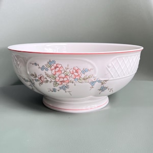 May include: White ceramic serving bowl with a pedestal base. The bowl features a floral design with pink and blue flowers, green leaves, and a red rim. The side has a textured diamond pattern.