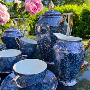 Puede incluir: Un juego de té que incluye una tetera, una jarra de leche y tazas con platillos, decorados con un esmalte moteado azul oscuro e iridiscente con asas y bordes dorados. El conjunto incluye una tetera con tapa, un azucarero y varias tazas a juego.