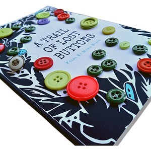May include: A book titled "A Trail of Lost Buttons" with poems by Maria Biren. The cover is black with white and blue floral designs and is decorated with colorful buttons.