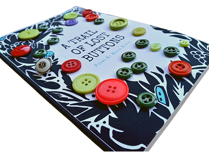 May include: A book titled "A Trail of Lost Buttons" with poems by Maria Biren. The cover is black with white and blue floral designs and is decorated with colorful buttons.