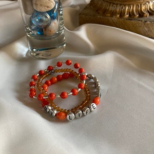 May include: Three beaded bracelets with orange and brown beads. The bracelets spell out "MY LIFE" in silver letters.