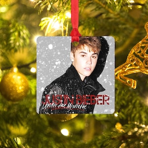 May include: Square Christmas ornament featuring a photo of Justin Bieber in a black leather jacket with a snowy background. The text "Justin Bieber Under the Mistletoe" is printed in red.