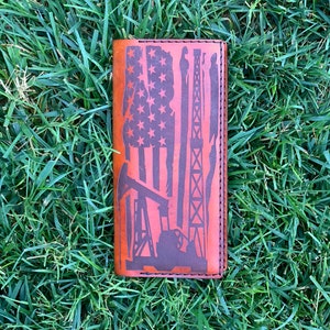 May include: A brown leather wallet featuring an American flag design and oil rig silhouettes. The wallet is rectangular with stitched edges. The flag design includes stars and stripes, and the oil rig silhouettes are in dark brown.