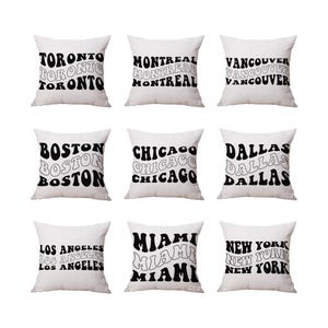 May include: Nine white throw pillows with black text featuring city names: Toronto, Montreal, Vancouver, Boston, Chicago, Dallas, Los Angeles, Miami, and New York. The text is stacked and in a retro font.