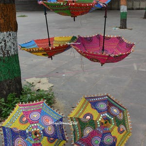 May include: Colorful, decorative umbrellas with intricate patterns and embellishments. Some are suspended from above, while others rest on the ground. The umbrellas feature a variety of colors, including pink, yellow, blue, and green, with decorative trim.