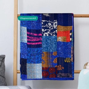 Könnte beinhalten: Eine blaue Patchwork-Decke mit verschiedenen bunten Mustern, darunter Blumen-, Paisley- und geometrische Designs. Die Decke hängt an einem Holzgestell.