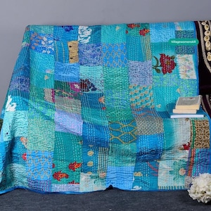May include: A turquoise blue patchwork quilt featuring a mix of patterns and colours, including floral designs, geometric shapes, and stripes. The quilt is folded and displayed on a sofa with a book and a bunch of white flowers.