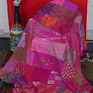May include: A pink and purple patchwork quilt made from recycled saris. The quilt is folded and draped over a chair. The quilt has a variety of patterns and textures, including floral, geometric, and paisley.