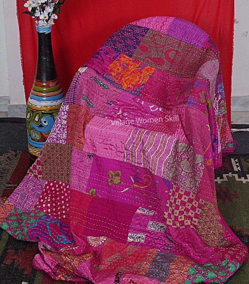May include: A pink and purple patchwork quilt made from recycled saris. The quilt is folded and draped over a chair. The quilt has a variety of patterns and textures, including floral, geometric, and paisley.