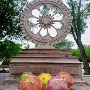 K&ouml;nnte beinhalten: Eine gro&szlig;e, aufwendig geschnitzte Steinskulptur eines Rades steht auf einer mehrstufigen Plattform. Davor sind mehrere farbenfrohe, offene Sonnenschirme mit goldenen Blumenmustern und Perlenkanten ausgestellt. Die Sonnenschirme sind in Gr&uuml;n-, Gelb-, Orange- und Rosat&ouml;nen gehalten.