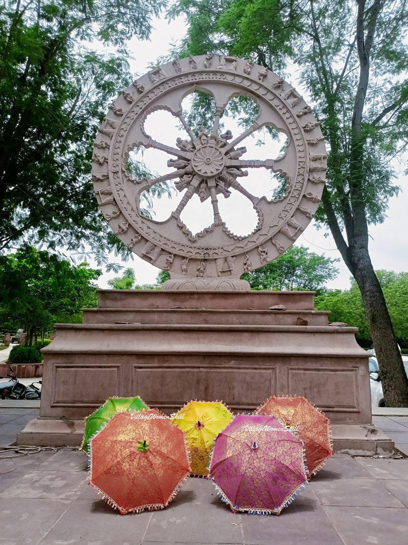 K&ouml;nnte beinhalten: Eine gro&szlig;e, aufwendig geschnitzte Steinskulptur eines Rades steht auf einer mehrstufigen Plattform. Davor sind mehrere farbenfrohe, offene Sonnenschirme mit goldenen Blumenmustern und Perlenkanten ausgestellt. Die Sonnenschirme sind in Gr&uuml;n-, Gelb-, Orange- und Rosat&ouml;nen gehalten.
