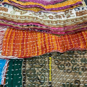 May include: A stack of colorful kantha quilts with intricate stitching and patterns. The quilts are made from recycled saris and are a traditional Indian craft. The top quilt is orange and red with white stitching, the next is brown and green with white stitching, and the bottom quilt is blue and green with white stitching. The quilts are all folded in half and are stacked on top of each other. The text "@VillageWomenSkill" is visible on the bottom right corner of the image.