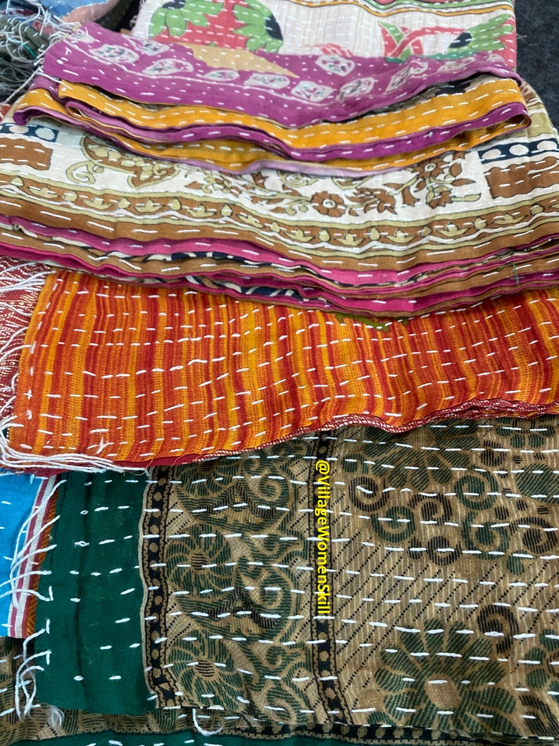 May include: A stack of colorful kantha quilts with intricate stitching and patterns. The quilts are made from recycled saris and are a traditional Indian craft. The top quilt is orange and red with white stitching, the next is brown and green with white stitching, and the bottom quilt is blue and green with white stitching. The quilts are all folded in half and are stacked on top of each other. The text "@VillageWomenSkill" is visible on the bottom right corner of the image.