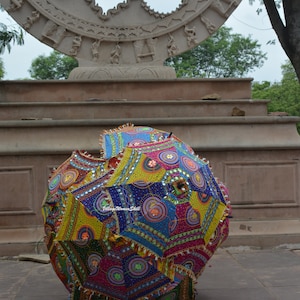 May include: A colorful, patterned umbrella with a patchwork design, featuring vibrant hues of blue, yellow, pink, and green. The umbrella has decorative tassels and is set against a stone backdrop.
