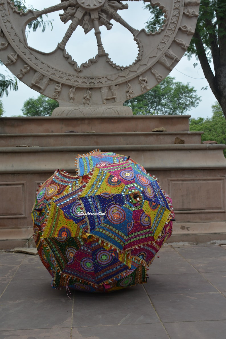 May include: A colorful, patterned umbrella with a patchwork design, featuring vibrant hues of blue, yellow, pink, and green. The umbrella has decorative tassels and is set against a stone backdrop.