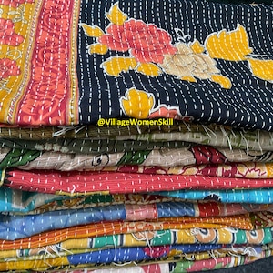 May include: A stack of colorful kantha quilts with floral patterns. The top quilt is black with yellow and red flowers. The quilts are hand-stitched and made from recycled saris. @VillageWomenSkill