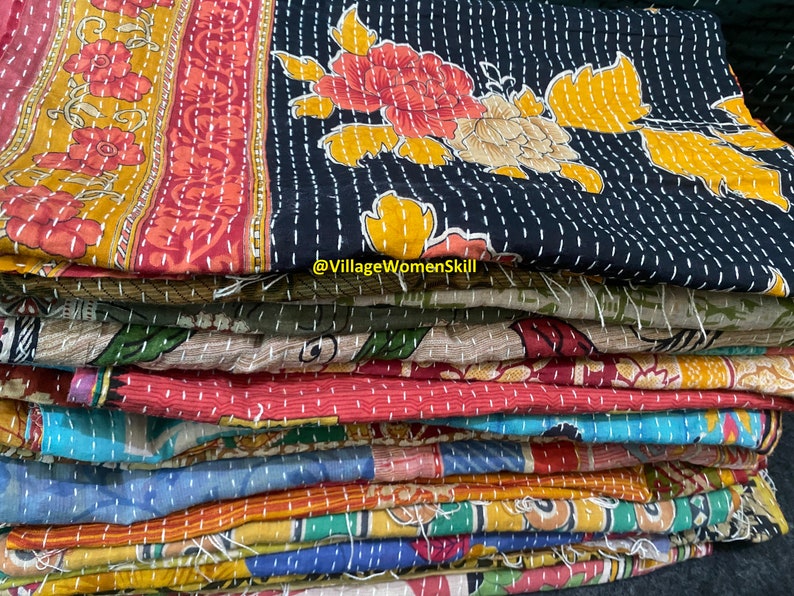 May include: A stack of colorful kantha quilts with floral patterns. The top quilt is black with yellow and red flowers. The quilts are hand-stitched and made from recycled saris. @VillageWomenSkill