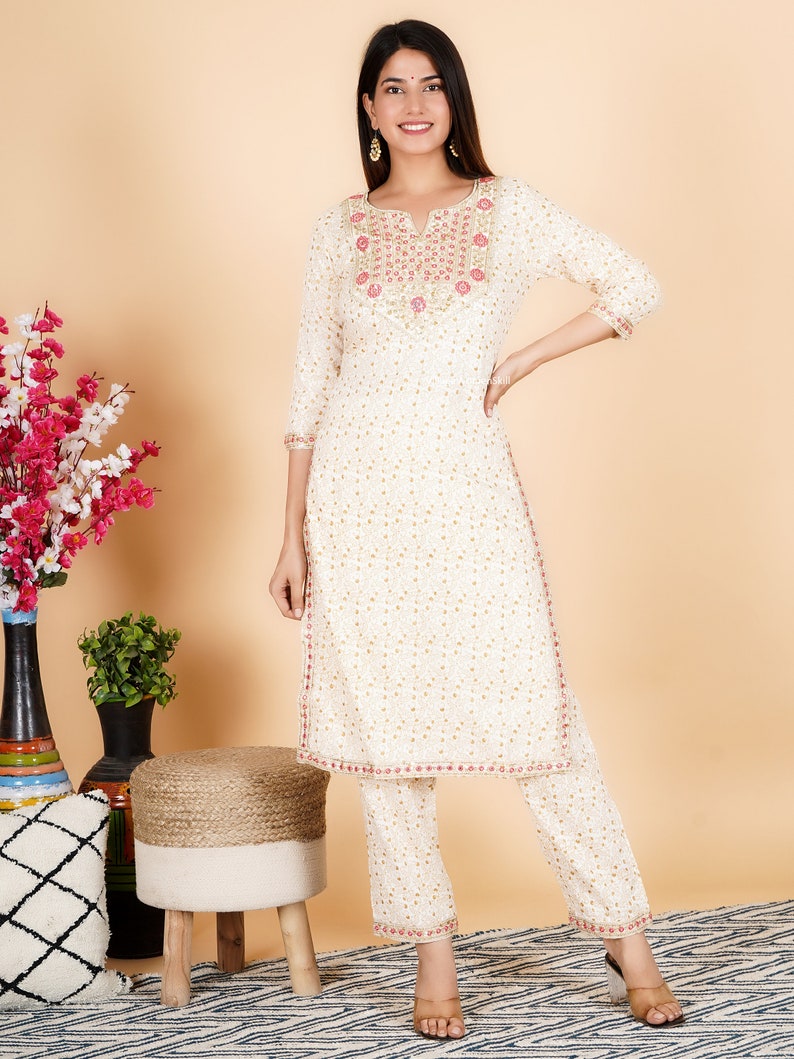 May include: White cotton kurta and palazzo set with gold and pink embroidery. The kurta has a V-neckline and three-quarter sleeves. The palazzo pants have a wide leg.