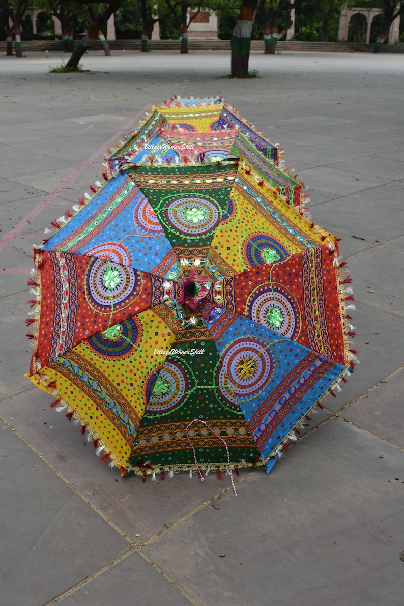 May include: A row of colorful, open decorative umbrellas with intricate patterns and embellishments. The umbrellas feature a patchwork design in red, yellow, blue, and green, with decorative tassels along the edges. The umbrellas are resting on a gray stone surface.