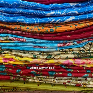 May include: A stack of colorful fabric scarves with floral and geometric patterns. The scarves are folded neatly and arranged in a pile. The text "Village Women Skill" is visible on the bottom of the pile.