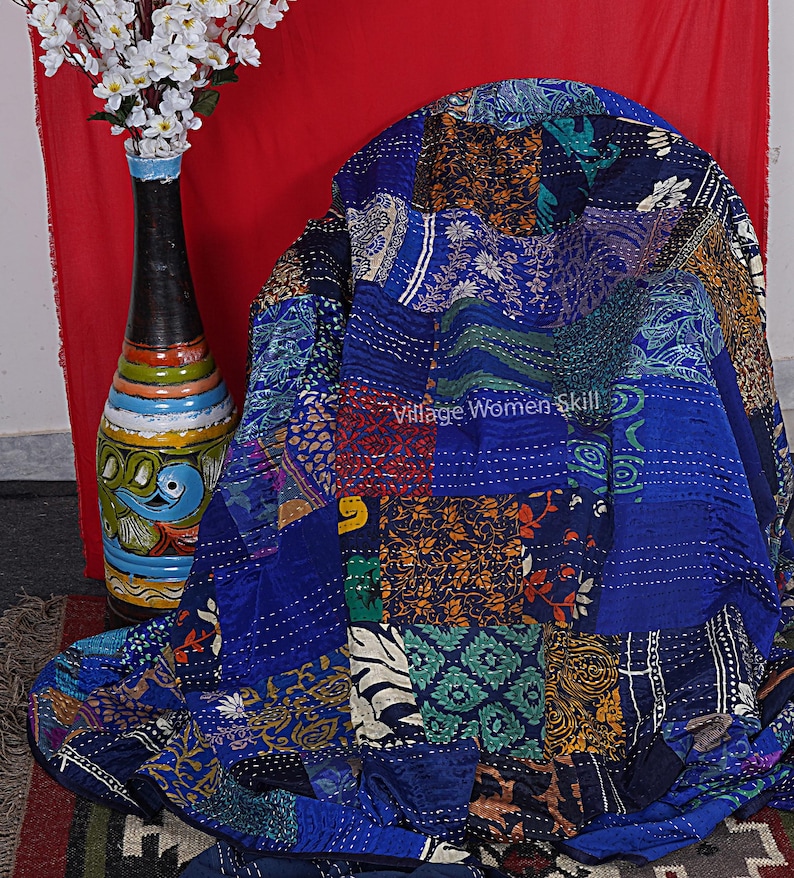 May include: A blue kantha quilt with a patchwork design featuring various floral and geometric patterns. The quilt is made of recycled saris and is hand-stitched. "Village Women Skill" is written on the quilt.