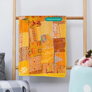 May include: A yellow, orange and brown patchwork quilt with a yellow border. The quilt is hanging on a wooden ladder. The text "Villagewomenskill" is visible on the quilt.