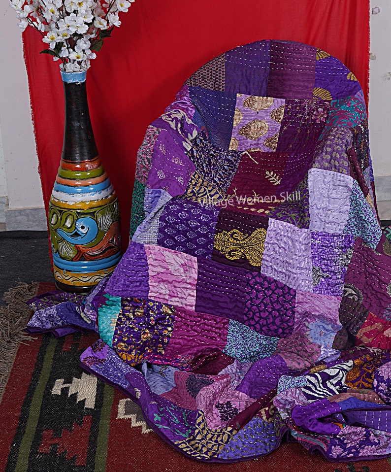 May include: A purple and pink patchwork quilt made from recycled saris. The quilt is folded and draped over a chair. The text "Village Women Skill" is visible on the quilt.