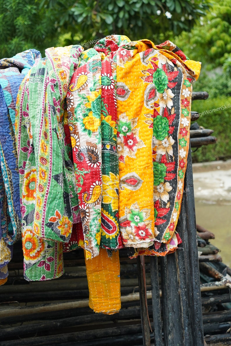 Puede incluir: Una pila de colchas kantha coloridas con motivos florales en tonos rojos, amarillos, verdes y azules. Las colchas est&aacute;n colgadas en un estante de madera.