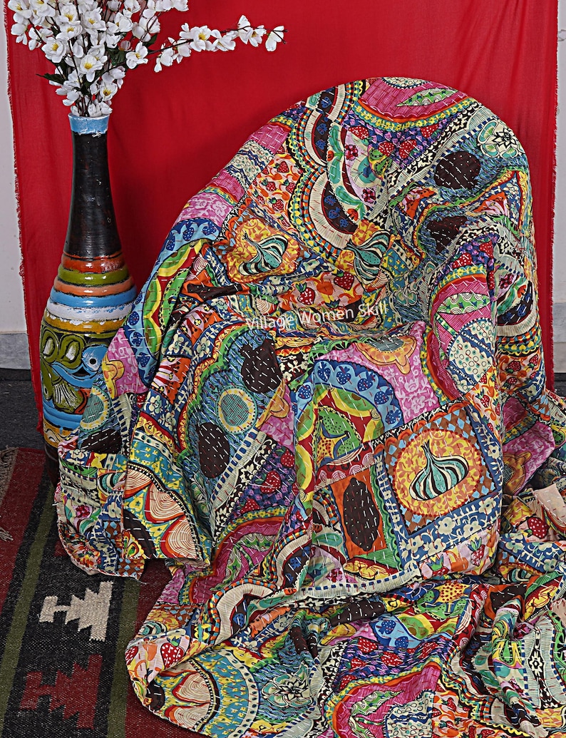 Puede incluir: Una colcha kantha colorida con un dise&ntilde;o de patchwork que presenta varios patrones y colores, incluyendo rosa, azul, verde, amarillo y marr&oacute;n. La colcha est&aacute; hecha de algod&oacute;n y est&aacute; cosida a mano. Est&aacute; doblada y drapeada sobre una silla.