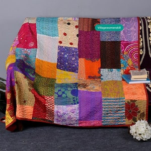 May include: A colorful patchwork quilt made from repurposed saris. The quilt is made up of squares of different colors and patterns, including pink, purple, orange, green, and blue. The quilt is folded and draped over a couch. The text "Villagewomenskill" is visible in the upper right corner of the image.