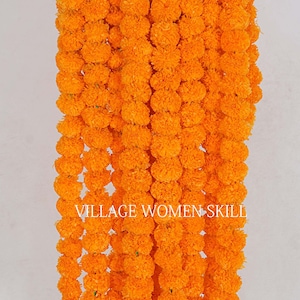 May include: A garland of bright orange marigold flowers, strung together in a long strand. The garland is hanging from a metal rod, and the ends of the garland are tied with green string. The text "VILLAGE WOMEN SKILL" is visible on the garland.
