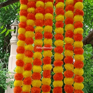 May include: A garland of yellow and orange paper flowers, arranged in alternating colors. The garland is made of multiple strands, creating a thick and full look. The garland is hanging from a tree branch.