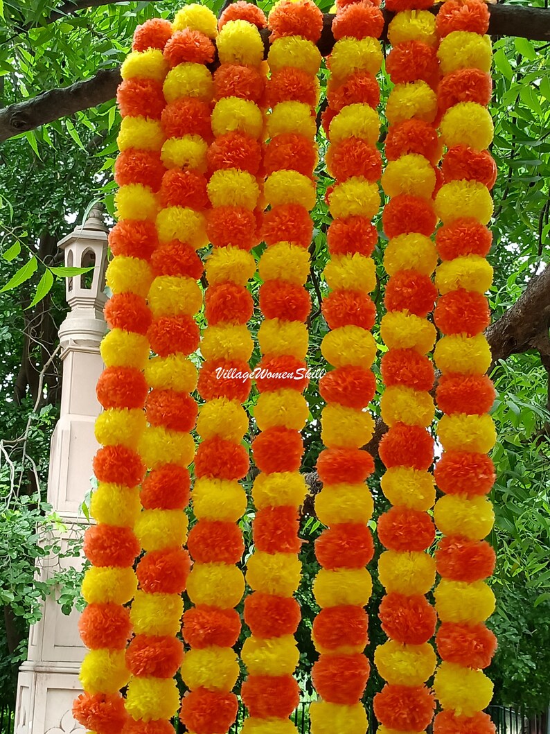 May include: A garland of yellow and orange paper flowers, arranged in alternating colors. The garland is made of multiple strands, creating a thick and full look. The garland is hanging from a tree branch.