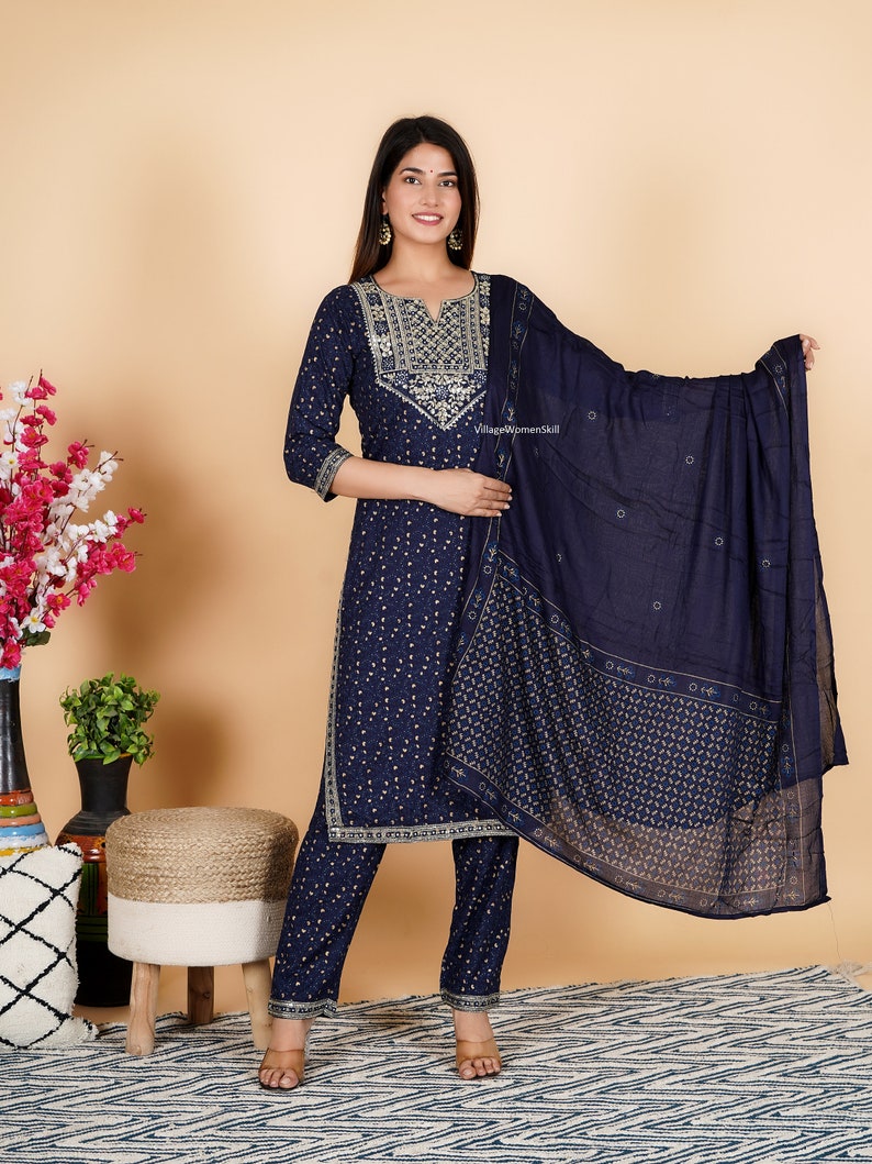 May include: A navy blue three-piece suit with gold embroidery. The suit includes a long-sleeved kurta, trousers, and a dupatta. The kurta has a V-neckline and a floral pattern. The trousers are straight-leg and the dupatta is sheer with a floral pattern. The text "Village Womenskill" is visible on the dupatta.