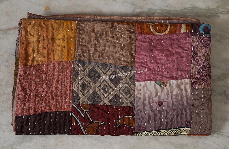 May include: A patchwork quilt made of various fabrics in shades of brown, pink, and orange. The quilt is hand-stitched with small, evenly spaced stitches. The quilt is folded in half and lying on a light-colored surface. The text "Village Women Skill" is visible in the center of the quilt.