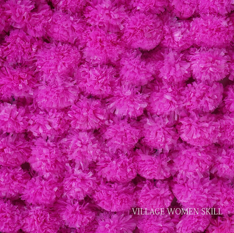 May include: A close-up of a wall covered in bright pink paper flowers. The flowers are made of tissue paper and are arranged in a random pattern. The text "VILLAGE WOMEN SKILL" is visible at the bottom of the image.
