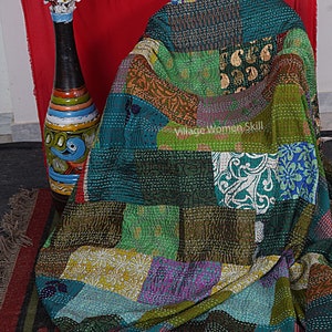 May include: A colorful patchwork quilt made from recycled saris. The quilt is a mix of green, blue, brown, and purple fabrics. The quilt is folded and draped over a chair. The text "Village Women Skill" is visible on the quilt.