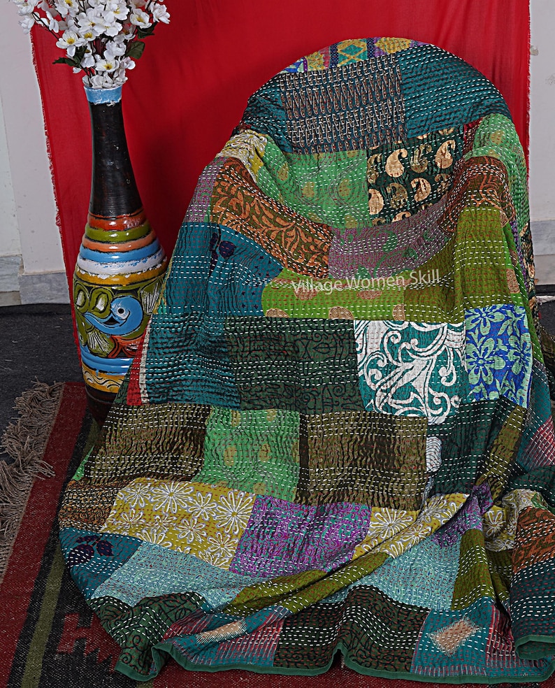 May include: A colorful patchwork quilt made from recycled saris. The quilt is a mix of green, blue, brown, and purple fabrics. The quilt is folded and draped over a chair. The text "Village Women Skill" is visible on the quilt.