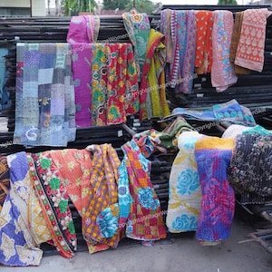 Puede incluir: Una pila de colchas kantha coloridas, hechas de saris reciclados, se exhiben en un estante de metal. Las colchas est&aacute;n hechas en una variedad de patrones y colores, incluyendo azul, verde, rojo, amarillo y naranja. Las colchas est&aacute;n todas cosidas a mano y tienen un aspecto vintage &uacute;nico. Copyright@VillageWomenSkill