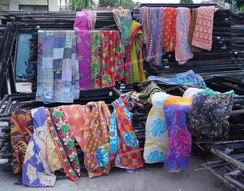 Puede incluir: Una pila de colchas kantha coloridas, hechas de saris reciclados, se exhiben en un estante de metal. Las colchas est&aacute;n hechas en una variedad de patrones y colores, incluyendo azul, verde, rojo, amarillo y naranja. Las colchas est&aacute;n todas cosidas a mano y tienen un aspecto vintage &uacute;nico. Copyright@VillageWomenSkill