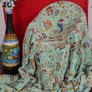 May include: A green kantha quilt with a floral pattern in shades of red, yellow, and blue. The quilt is folded and draped over a chair. The text "Village Women Skill" is visible on the quilt.