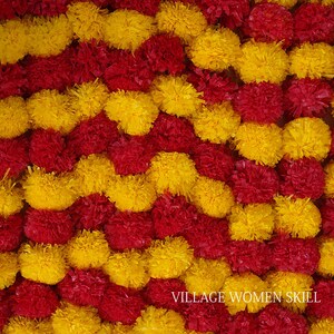May include: A close-up of a decorative garland made of red and yellow fabric flowers. The garland is arranged in a wavy pattern. The text "VILLAGE WOMEN SKILL" is visible at the bottom of the image.